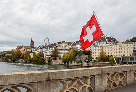 Swiss flag on a bridge in front of waterfront with old buildings, emblematic of the client Avenir Suisse - n c ag