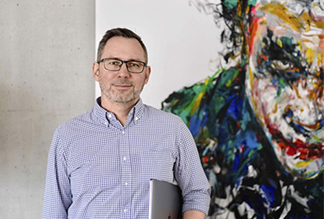 Thomas Kreindl steht mit Notebook im Arm vor einem modernen Gemälde - n c ag